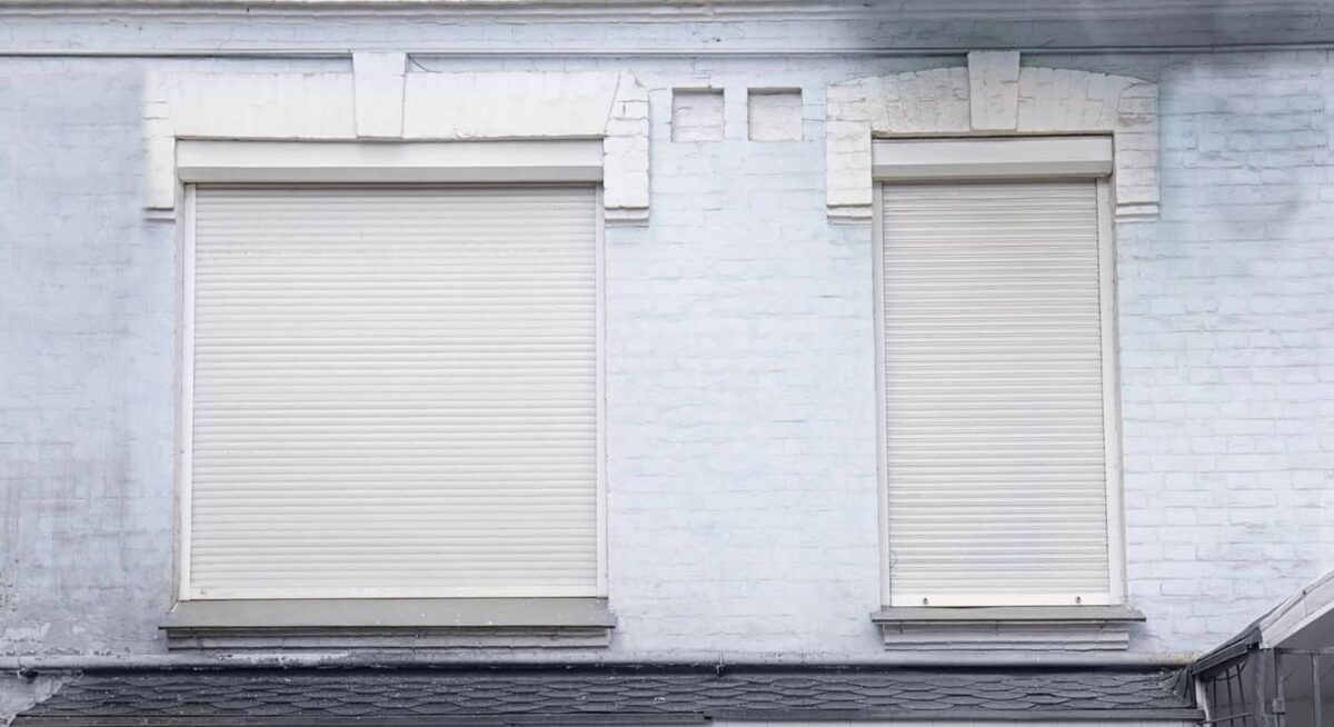 Deux volets roulants blancs fermés sur une façade de brique peinte en clair