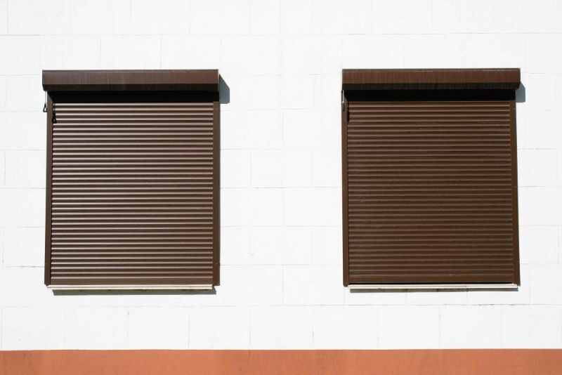 Deux volets roulants bruns fermés, leurs coffres en applique posés sous le linteau d'une façade claire
