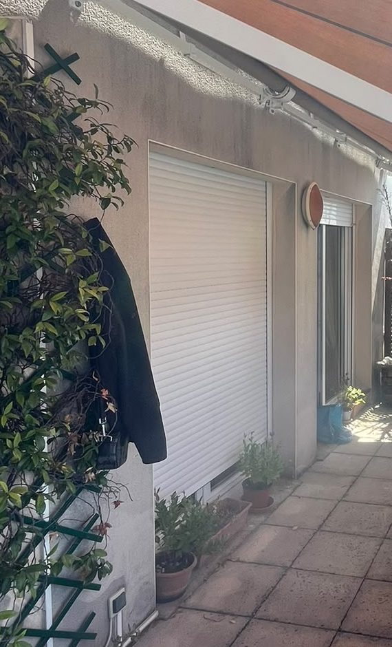 Volet roulant blanc fermé sur la porte-fenêtre d'un balcon filant à Paris 1er, façade en crépi, plantes en pots au pied du mur